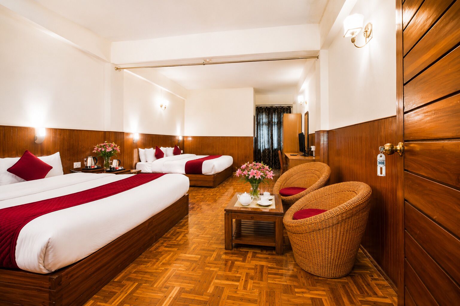 A hotel room with two beds, a wooden floor, a small table with a vase of flowers, and wicker chairs.