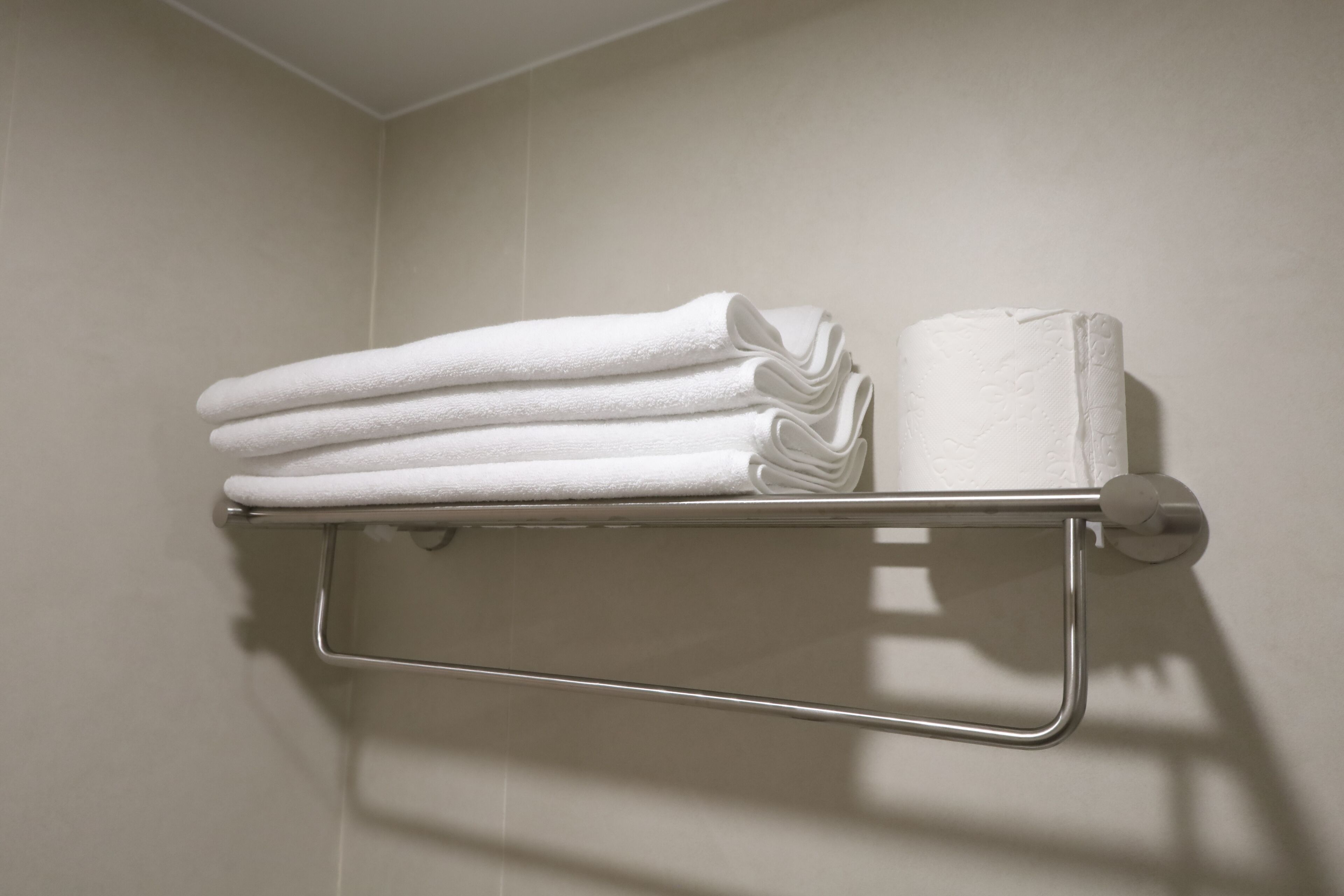 A metal towel rack with folded white towels and a roll of toilet paper against a plain wall.
