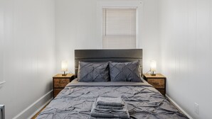 A neatly made bed with a grey duvet, two bedside tables with lamps, and a window with blinds.