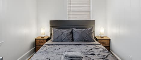 Una cama bien hecha con un edredón gris, dos mesitas de noche con lámparas y una ventana con persianas.