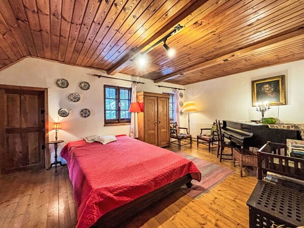 A cozy room with a wooden ceiling, a bed with a red blanket, a piano, and a desk with a lamp.
