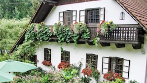 Un edificio blanco con múltiples ventanas, algunas con contraventanas, y un balcón con plantas colgantes. Hay una sombrilla verde y una silla en primer plano.