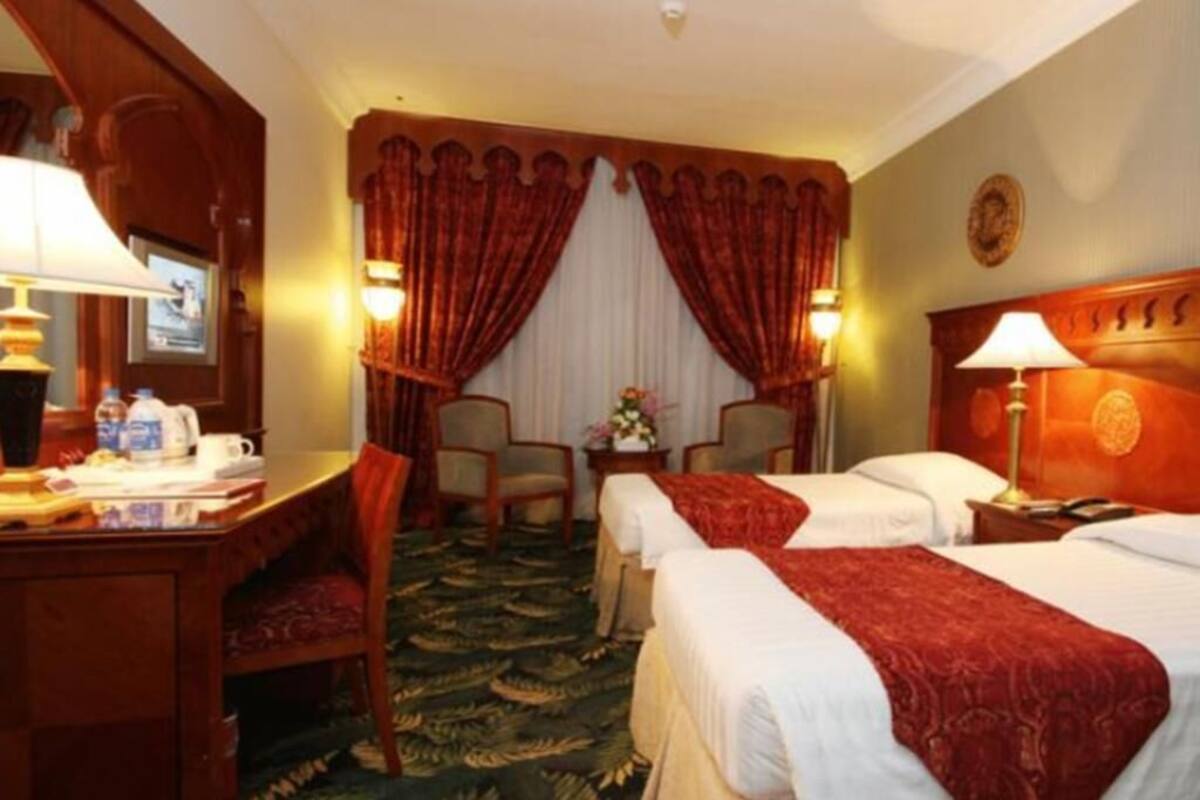 Habitación de hotel con dos camas, un escritorio con lámpara, una silla, una mesita con un florero y un ventanal con cortinas.