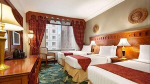 A hotel room with two beds, a wooden headboard, a desk with a lamp, a chair, and a view of buildings through large windows.
