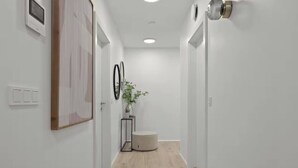 Un corridoio stretto con pareti bianche, pavimento in legno e una piccola zona salotto rotonda. Sono presenti luci a parete e un quadro incorniciato a sinistra.