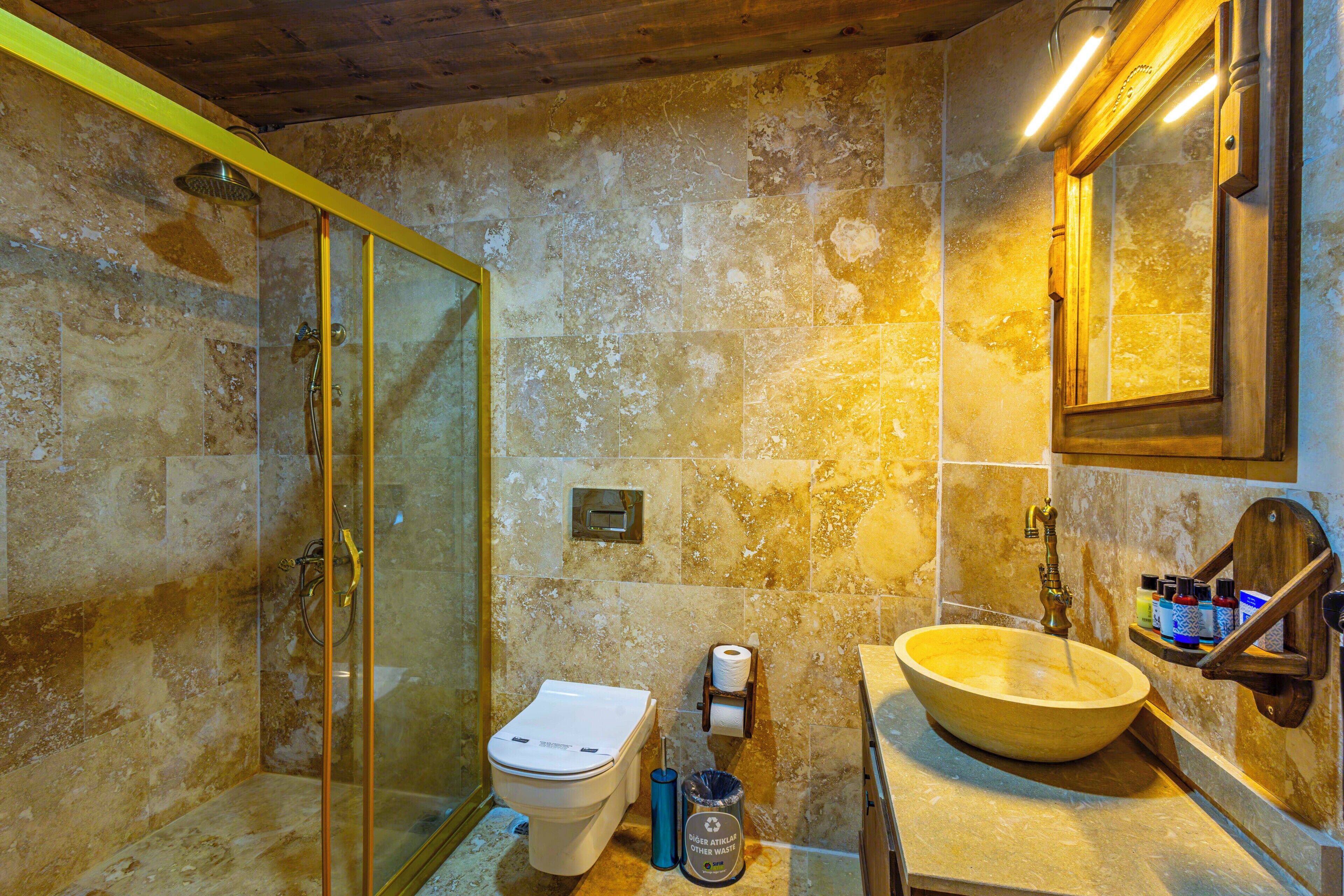 Un baño con ducha de vidrio, inodoro, lavamanos de madera y pared con azulejos de mármol.