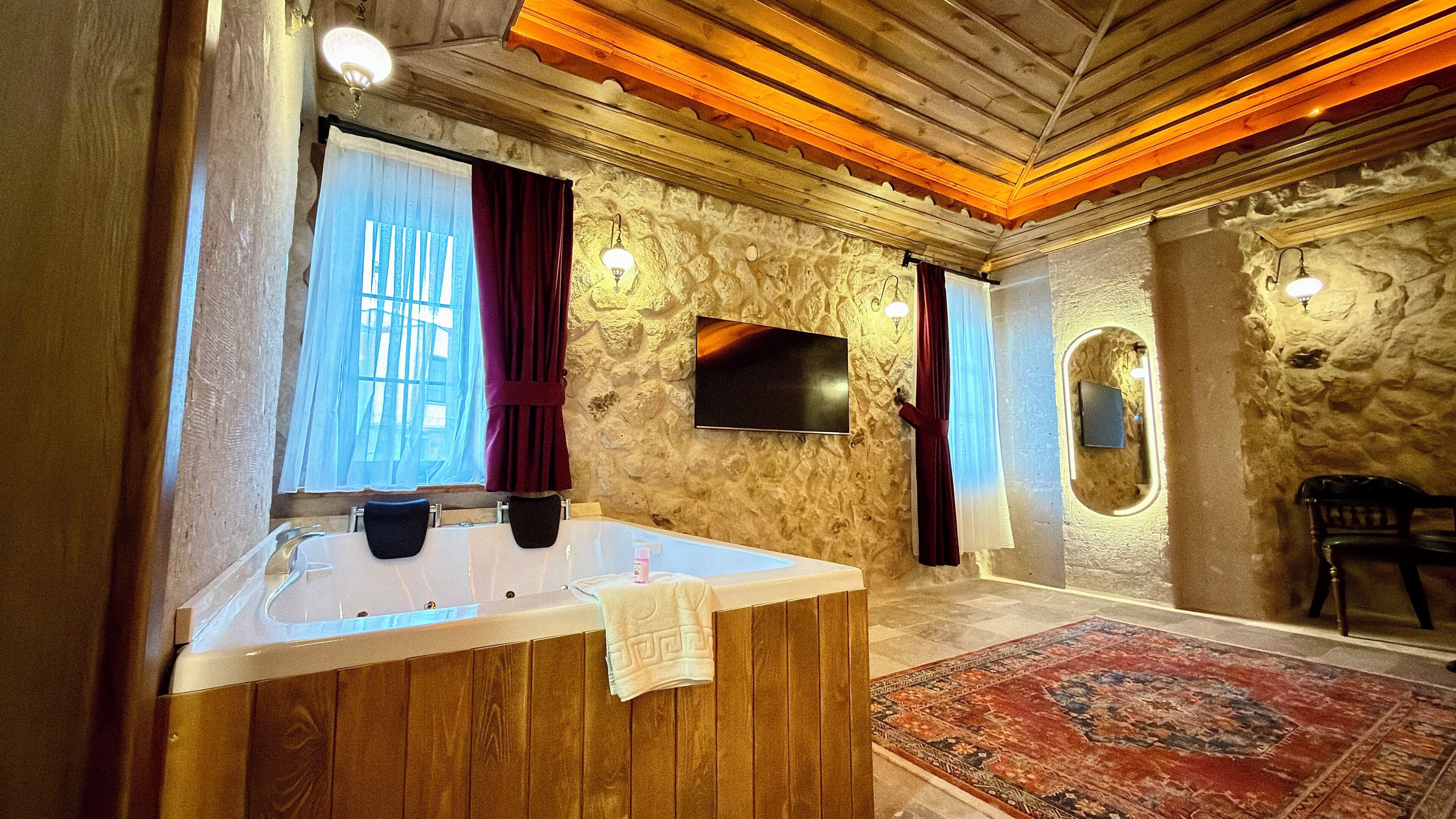 A bathroom with a bathtub, a wooden vanity, a stone wall, a television, and a mirror.