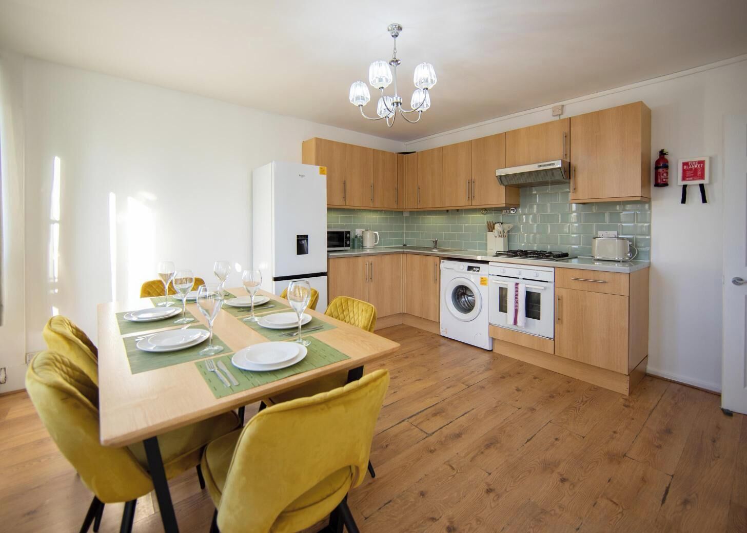 A modern kitchen with wooden cabinets, a built-in washing machine, a stove, and a dining area with yellow chairs and a table set for four.