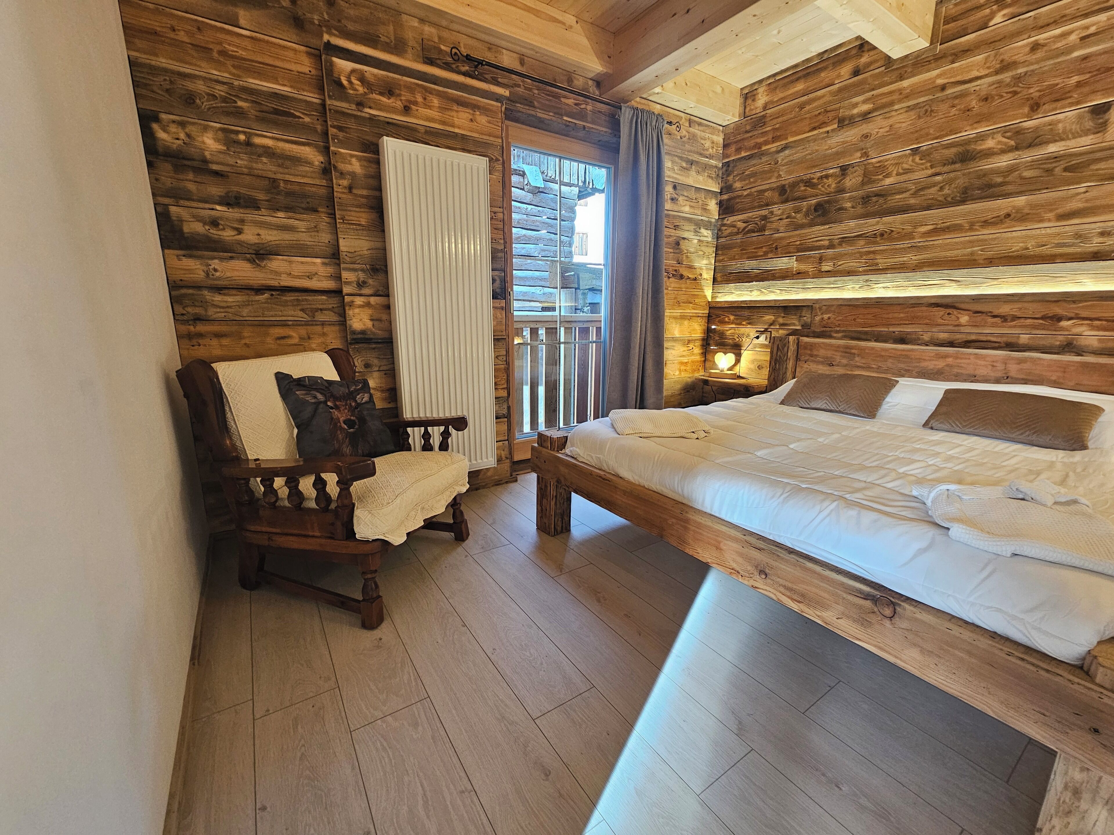 Une pièce de cabane en bois comprenant un lit, une chaise avec un coussin à motif de cerf, un radiateur et une fenêtre avec des rideaux.