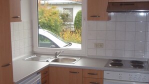 Cocina compacta con armarios de madera, lavavajillas blanco, estufa y ventana con vista a un coche y árboles.