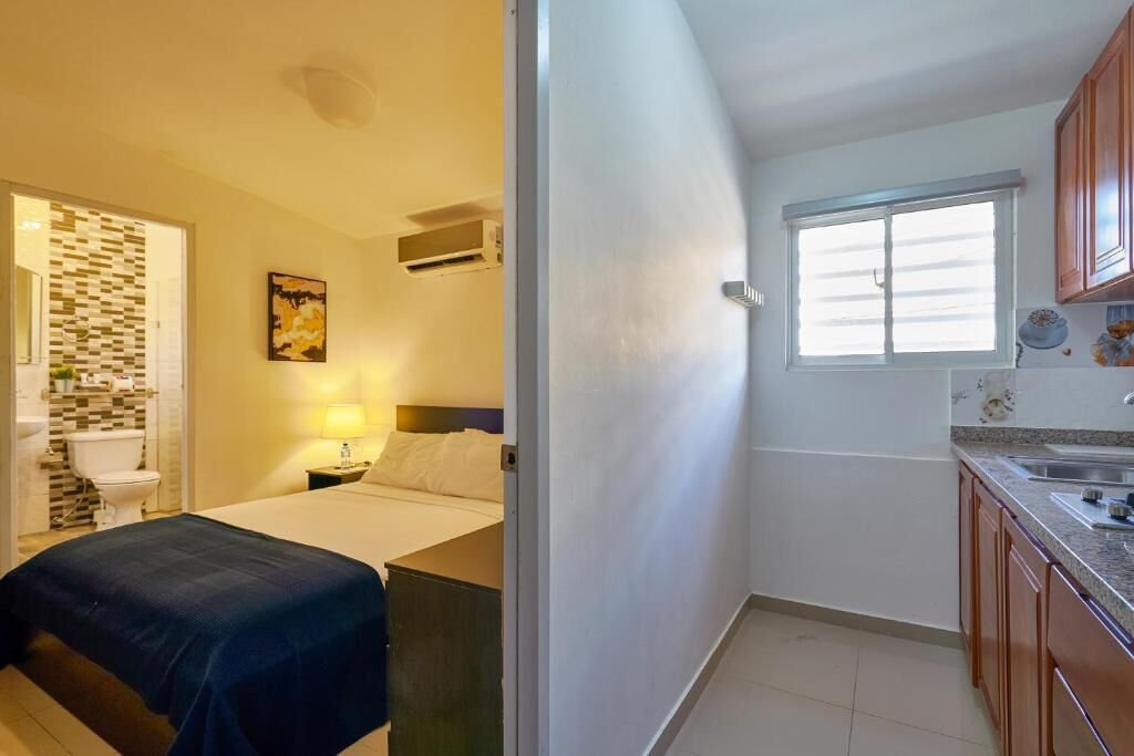 Habitación de hotel pequeña con cama, mesita de noche, lámpara y una kitchenette que incluye pileta y alacenas.
