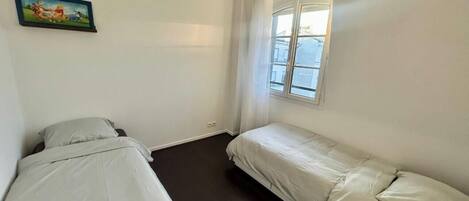 Un apartamento pequeño de una habitación con dos camas, una ventana con cortinas e imagen colgada en la pared.