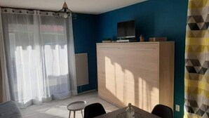 Une pièce comprenant un lit, une table agrémentée d’un objet décoratif, un téléviseur posé sur un meuble et une fenêtre ornée de rideaux légers.