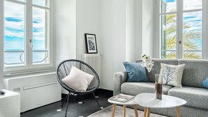 A modern living room with a grey sofa, a black chair, and a white coffee table. There is a framed picture on the wall and a radiator below it.