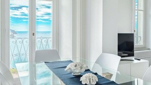 A modern dining area with a glass table, white chairs, and a view of the sea through large windows.