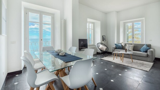 A spacious living area with a glass dining table, white chairs, a grey sofa, and a view of the sea.