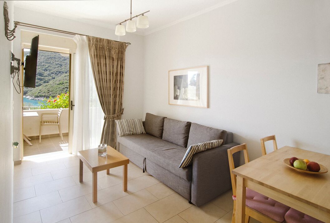 A living room with a sofa, a wooden table, and a dining table with fruit. There is a window with curtains, a framed picture on the wall, and a view of a hillside.