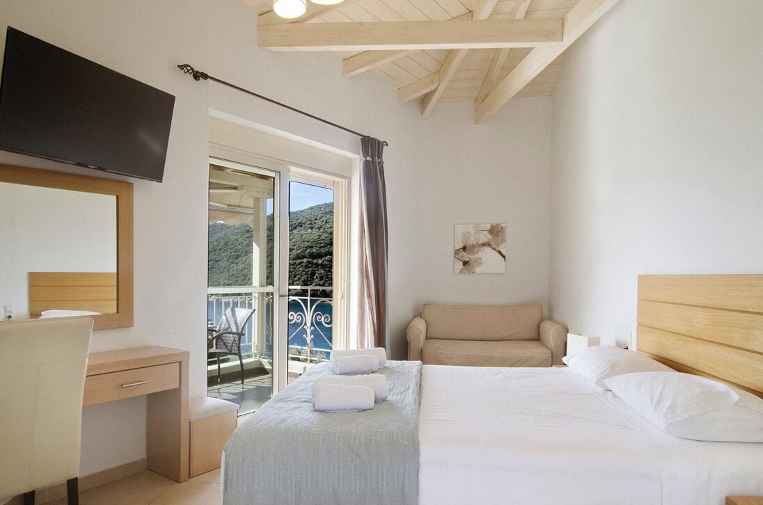 Ein Schlafzimmer mit Bett, Schreibtisch, Stuhl, Fernseher und einem Balkon mit Blick auf Grünflächen.