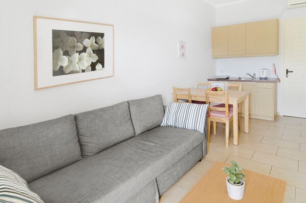 A compact living space with a grey sofa, a dining table with chairs, a small kitchen area, and a framed picture on the wall. - ROUDA VILLAGE (Lefkada)