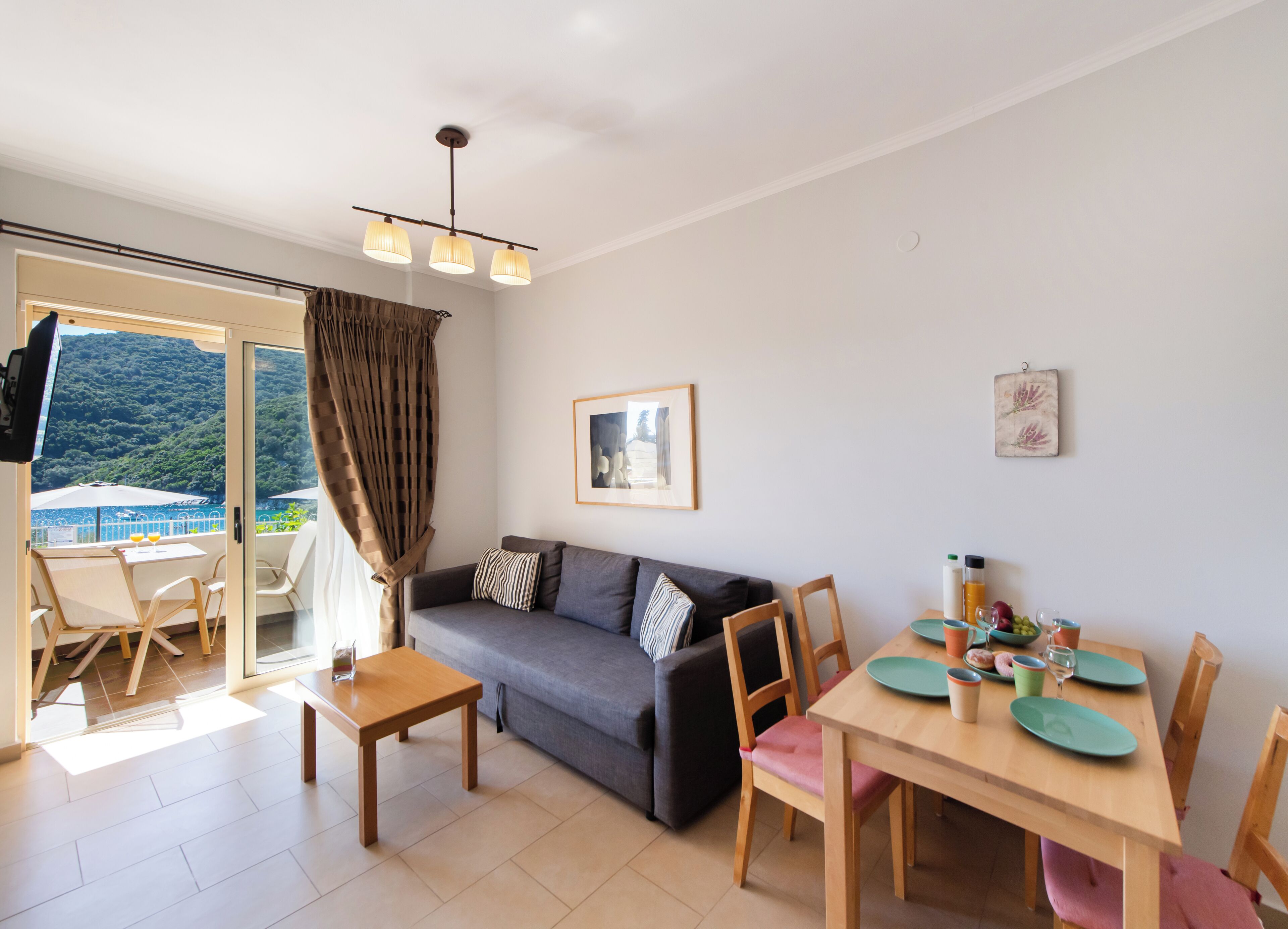 A living room with a sofa, a dining table with chairs, a TV, and a view of a balcony with a table and chairs.