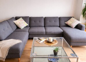 A grey sectional sofa with patterned cushions, a glass coffee table with books and a candle, and a small potted plant.