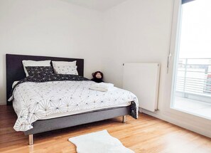 A modern bedroom with a grey bed, white and black patterned bedding, a small monkey plush toy, a white rug, and a window with a view of buildings.