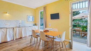 Ein helles, gelbes Zimmer mit einem Holzespeisetisch und -stühlen, einer Küchenzeile mit Spüle und Arbeitsplatte sowie einem an der Wand montierten Fernseher.