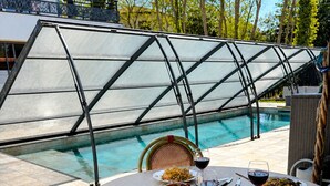 Une table ronde avec un plateau en verre, entourée de chaises, près d’une piscine dont une partie est protégée par une paroi en verre.