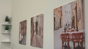 A hallway with three paintings depicting street scenes, a white shelf with a plant, and a white wall. - Domus Tiziano: Elegant Living Room 30m from Metro C - Center of Rome (Roma)