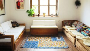 A cozy living room with a wooden bench, a white sofa, and a brown sofa with colorful cushions. There is a window with plants on the sill and a wall-mounted decoration. - Artist house with garden sauna in London (London)