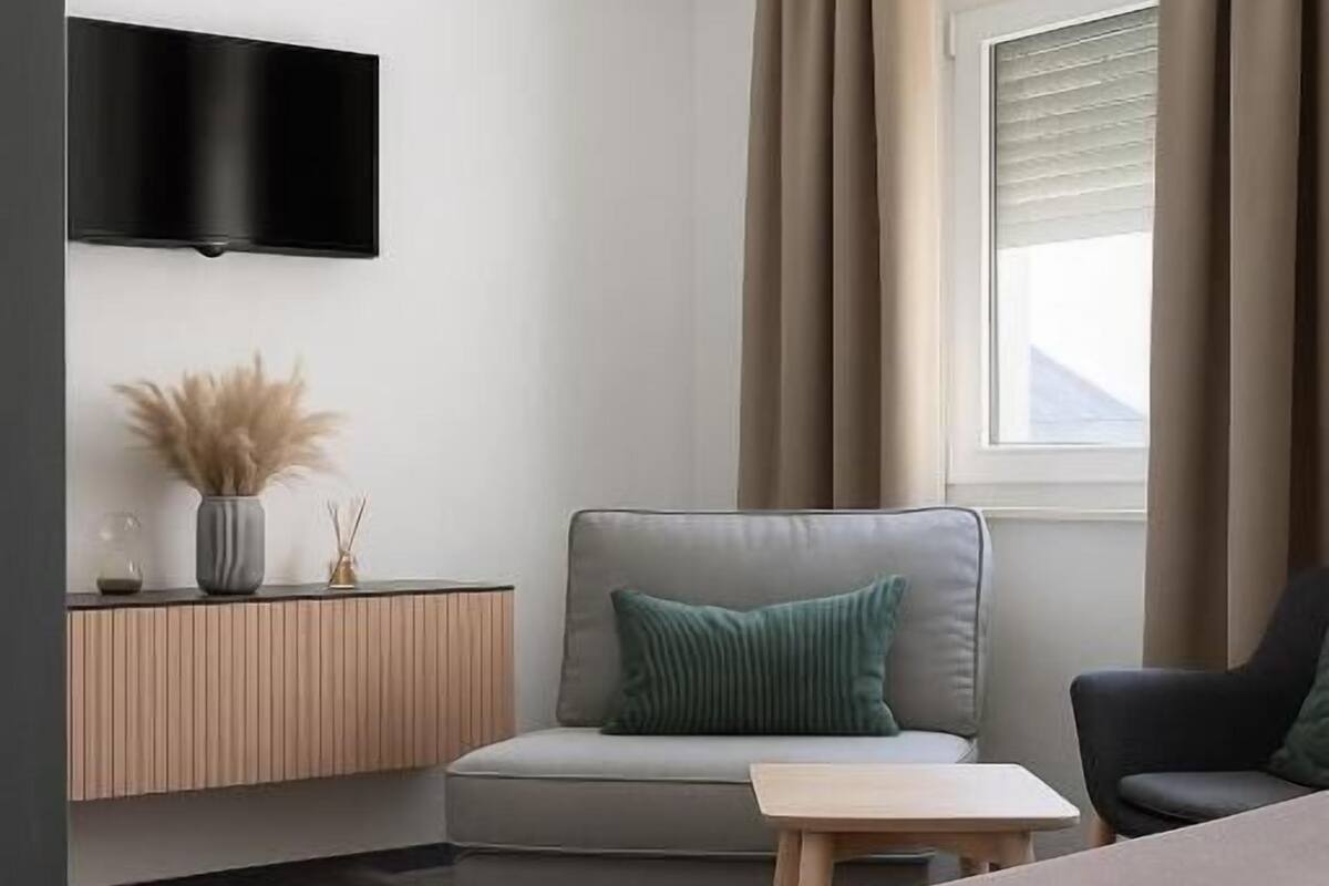 Een moderne hotelkamer met een bed, een zithoek met een bank en fauteuil, een klein tafeltje, een aan de muur bevestigde televisie en een raam met gordijnen.