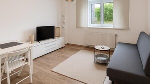 Een moderne woonkamer met een flatscreen televisie, een bank, een klein rond tafeltje en een raam met jaloezieën.