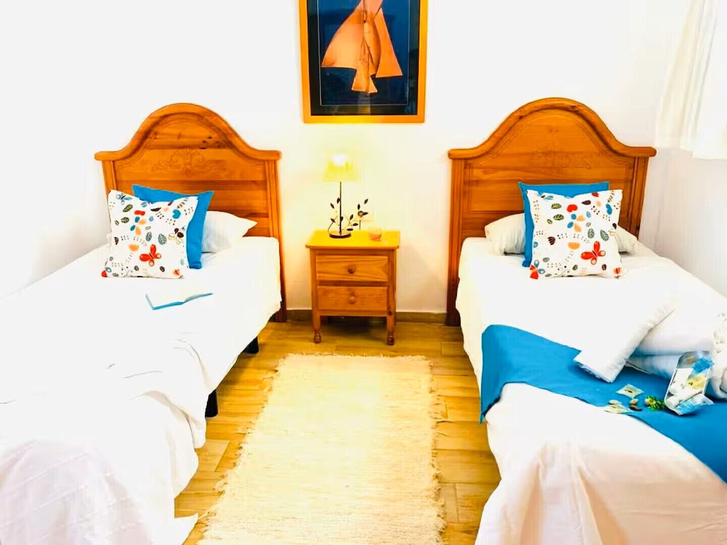 A double room with two wooden beds, each with a colorful pillow and a small bedside table with a lamp.