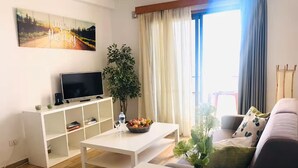 Een woonkamer met een televisie, witte meubels, een eettafel en een groot raam met gordijnen.
