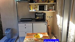 A compact kitchenette with a microwave, toaster, and a small oven. There is a trash bin and a table with a bowl. The room has a simple layout with a window and a door. - 40m², 100m from beach, garden, 1 bedroom, living room (sofa bed), fiber box, air conditioning, no pets (La Ciotat)