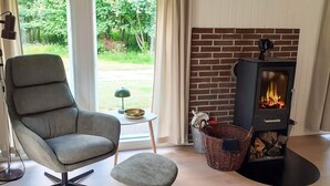 Un salon moderne avec un fauteuil gris, un petit tabouret, une cheminée et une vue sur la verdure à l’extérieur.