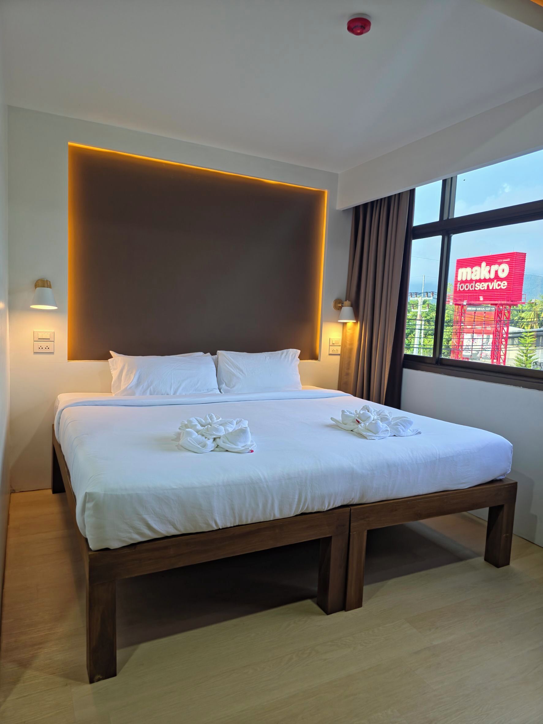 Quarto de hotel com cama grande, luminárias laterais, vista para o serviço de alimentação Makro e uma janela com cortinas.