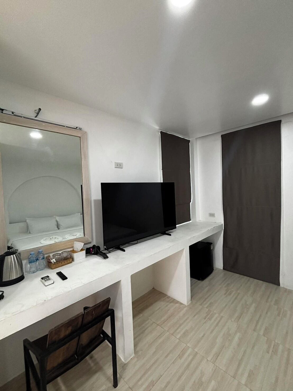 A modern hotel room with a white desk, a flat-screen TV, a mirror, a bed with white bedding, and a dark curtain.