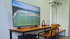 A room with a wooden desk, a chair, a television, a small refrigerator, a kettle, and a water bottle on the desk.