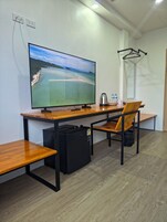 A room with a wooden desk, a chair, a television, a small refrigerator, a kettle, and a water bottle on the desk.