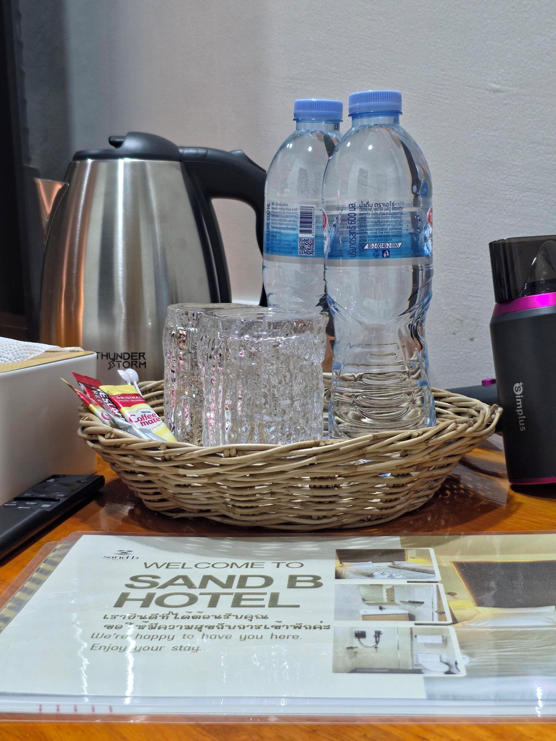 Un felpudo, una bolsita de café, un hervidor, dos botellas de agua, un vaso y una taza negra sobre una mesa.