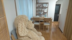 A room with a large plush toy on a chair, a dining table with chairs, a shelf with decorative items, and a door.