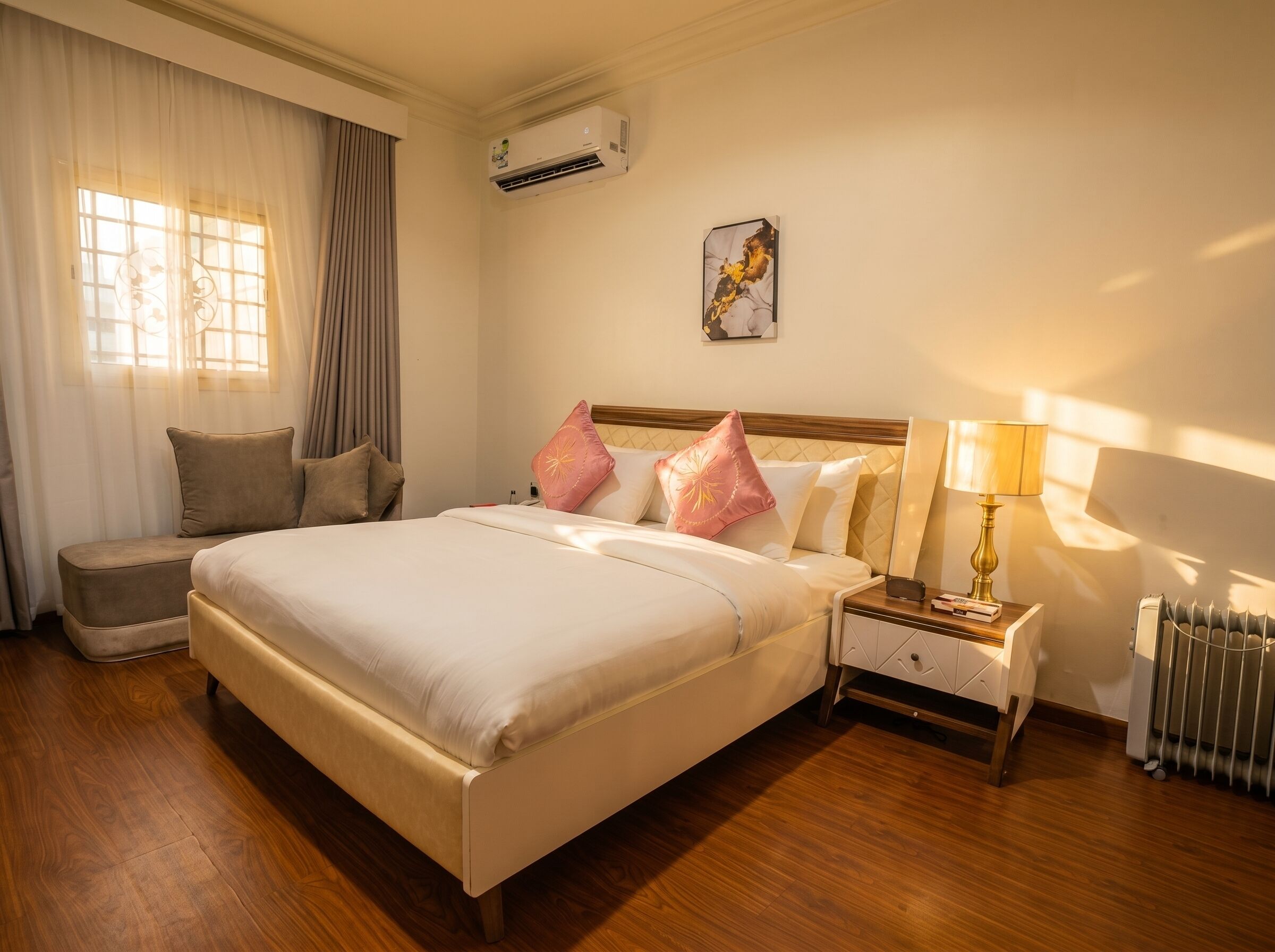 A hotel room with a bed, bedside table, lamp, radiator, and an air conditioning unit.