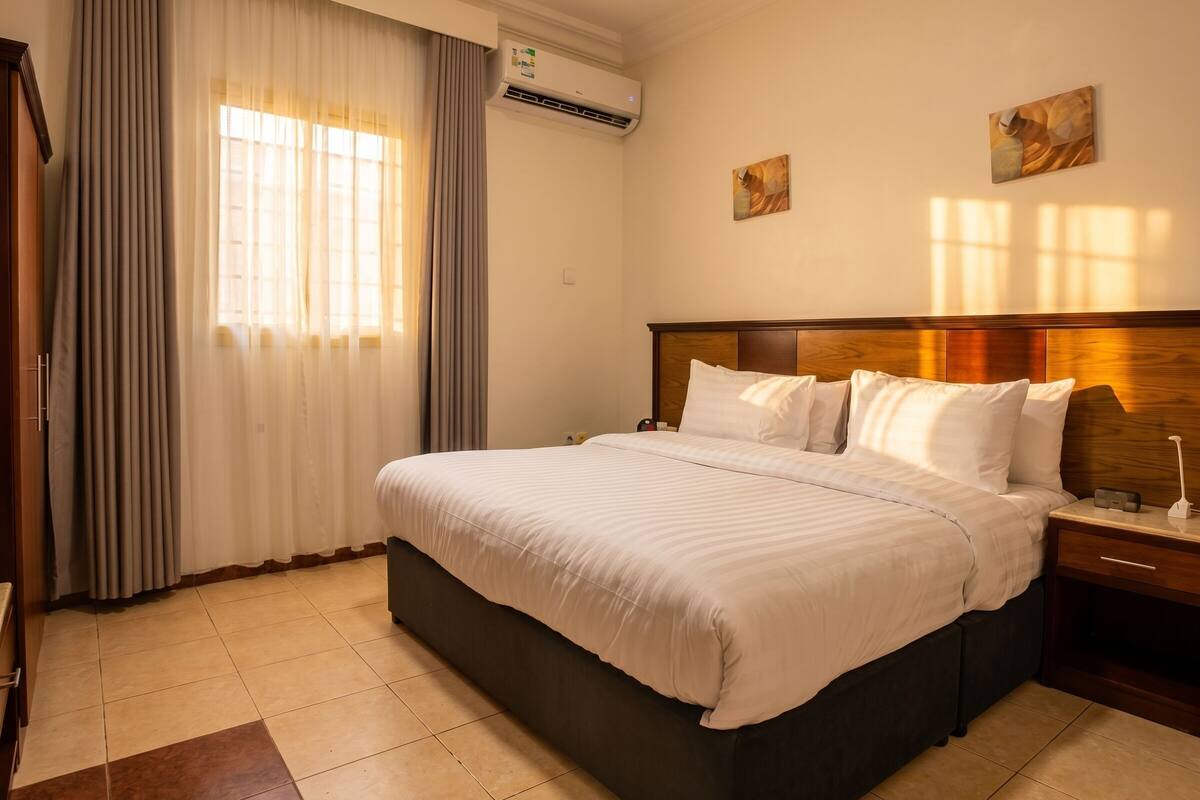 Ein Hotelzimmer mit Bett, Vorhängen, einer Klimaanlage, einem Nachttisch und Bildern an der Wand.