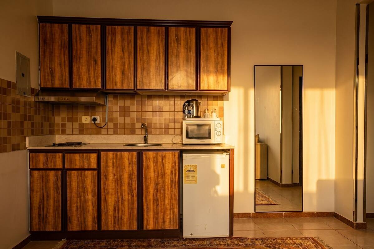 Eine kleine Küche mit Holzschränken, einer Mikrowelle, einem kleinen Kühlschrank und einer gefliesten Spritzschutzwand.