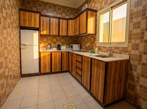 A compact kitchen with wooden cabinets, a refrigerator, a microwave, and a window. - Vinas Riyadh Serviced Apartments (Riyadh)