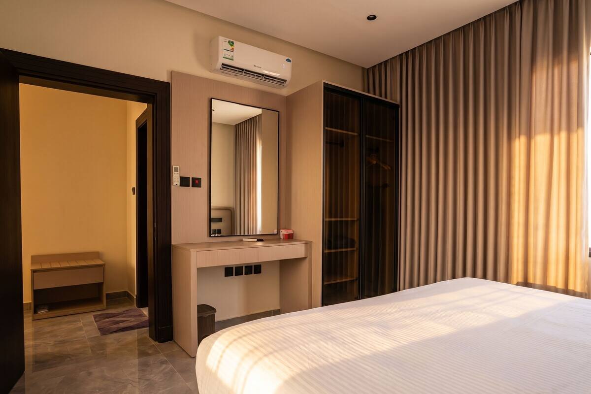 Ein Hotelzimmer mit Bett, Schminktisch mit Spiegel, integrierter Klimaanlage, Kleiderschrank und einer Tür zu einem weiteren Raum.