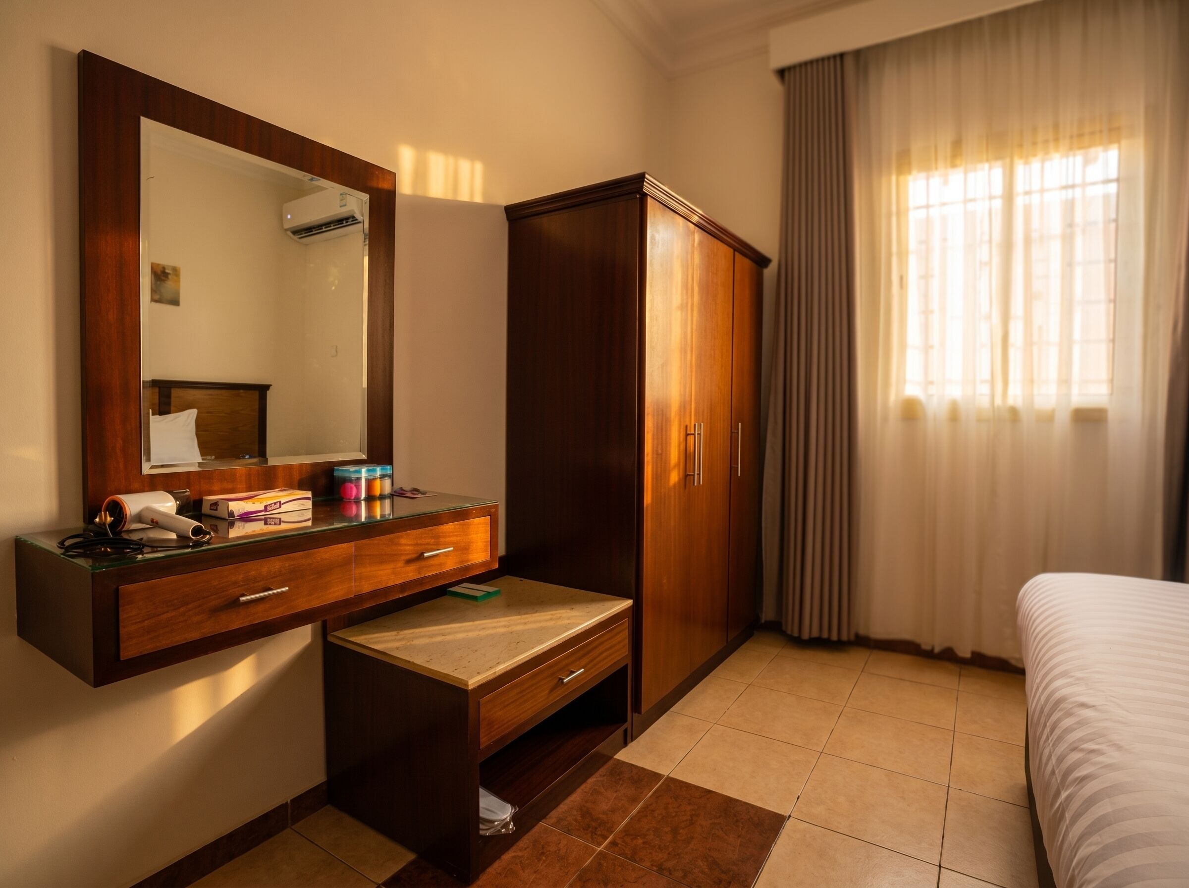 Quarto de hotel com guarda-roupa de madeira, espelho, mesa de cabeceira com abajur e uma cama.