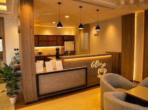 A modern coffee shop interior with a wooden reception desk, pendant lighting, and a seating area with a sofa and table. - Vinas Riyadh Serviced Apartments (Riyadh)