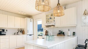 Una cocina moderna con armarios blancos, una isla central, lámparas colgantes de mimbre y vistas al exterior.